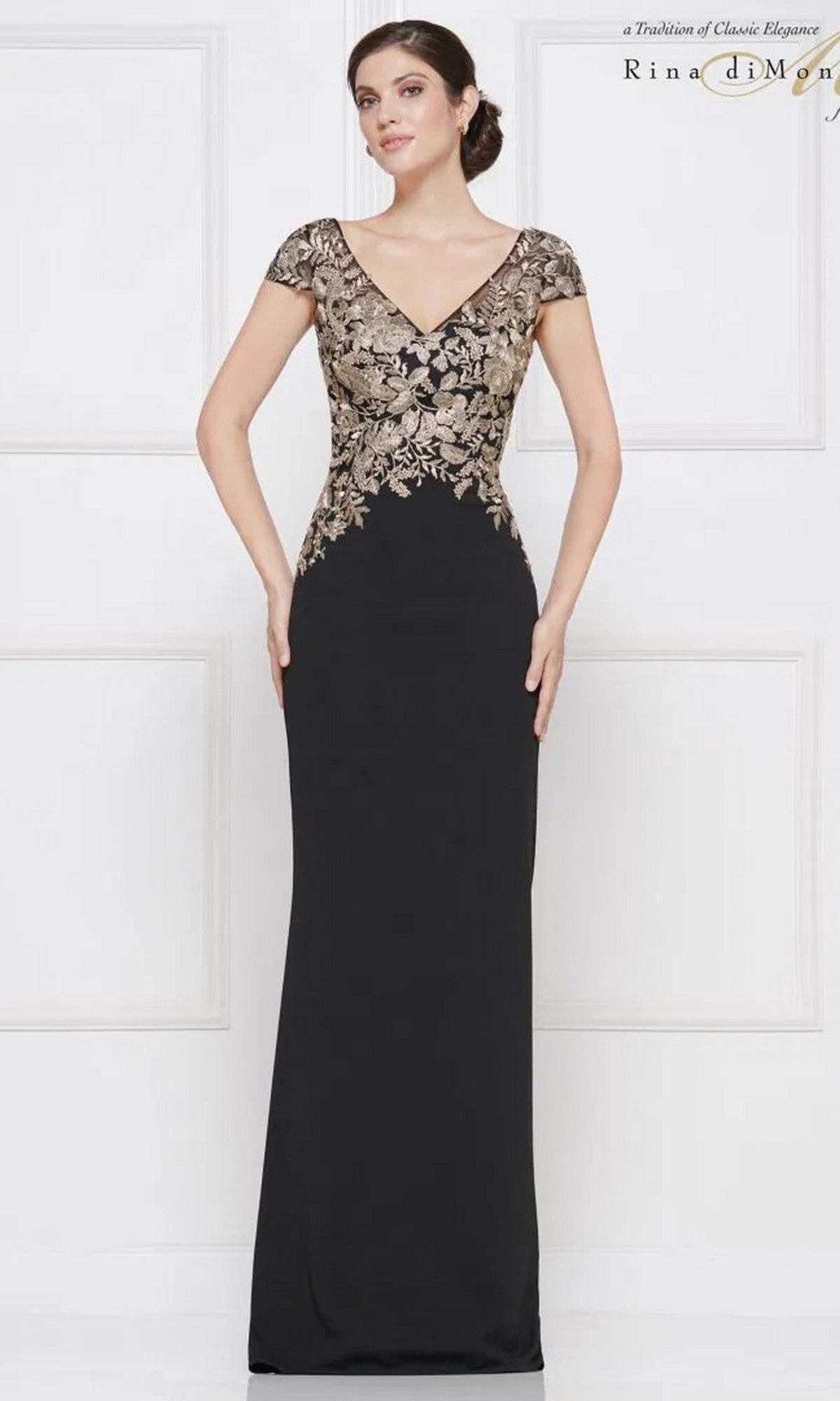 A woman models the Rina Di Montella RD2652, a long formal dress with short sleeves, gold-embroidered floral bodice details, and a sleek sheath skirt, standing before a white paneled wall.